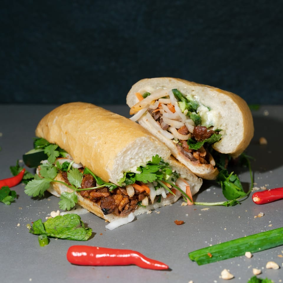 Two halves of a Saigon Blvd Bánh Mì sandwich filled with meat and vegetables, surrounded by chilies and herbs.