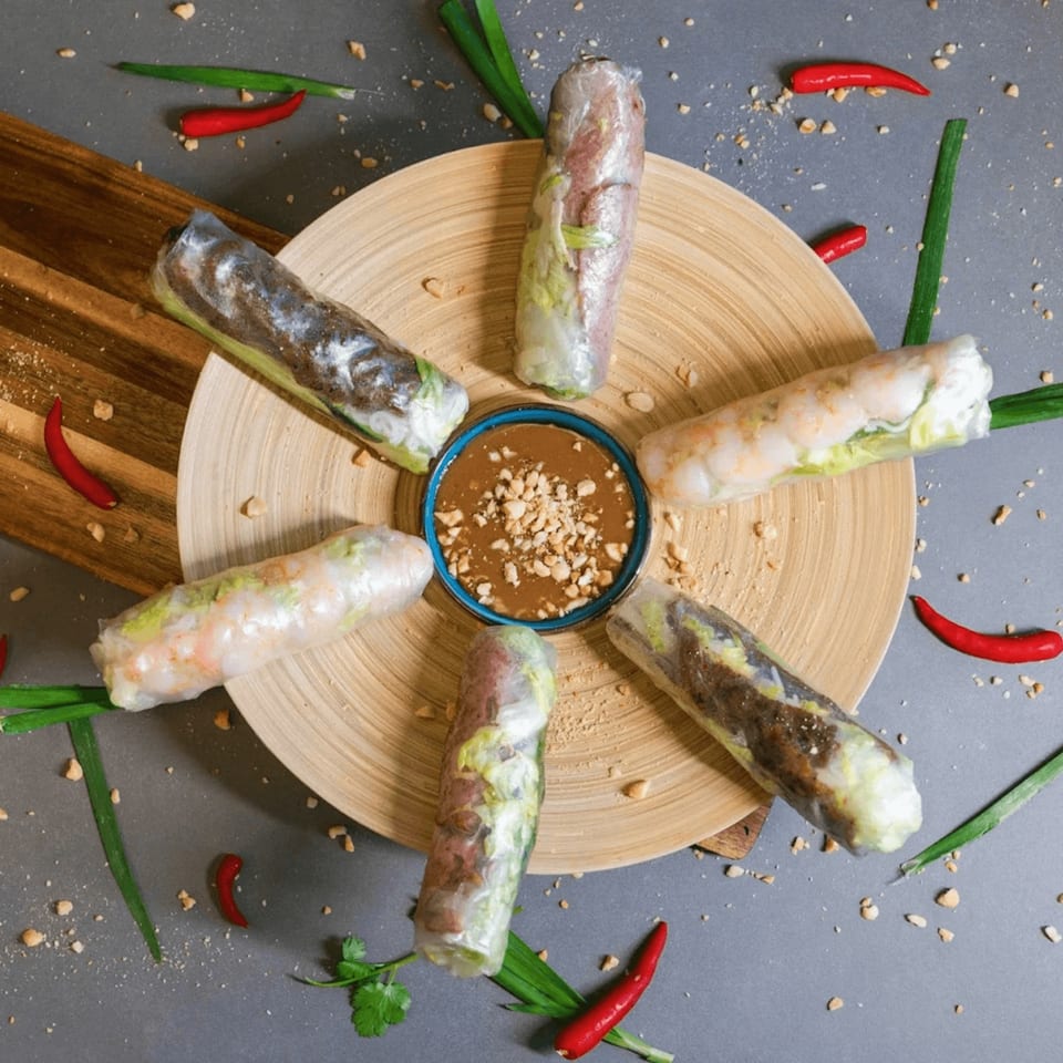 Six Vietnamese spring rolls with peanut dipping sauce on a bamboo plate, garnished with chilies and scallions.