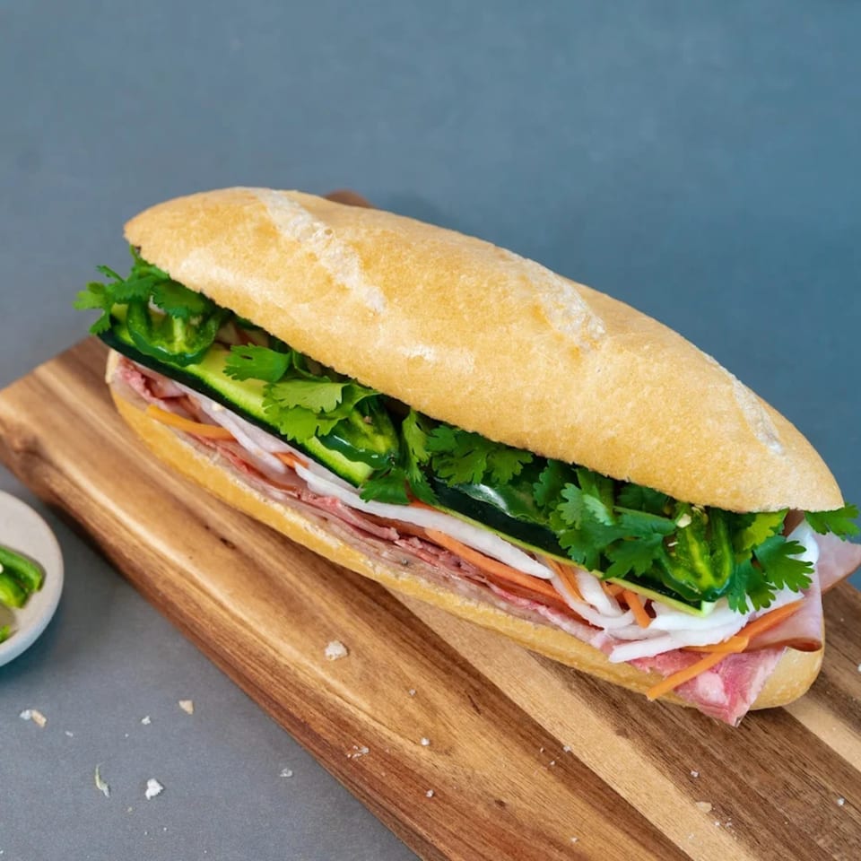 A Saigon Blvd Bánh Mì sandwich, filled with meat, pickled vegetables, cilantro, and jalapeños on a wooden board.