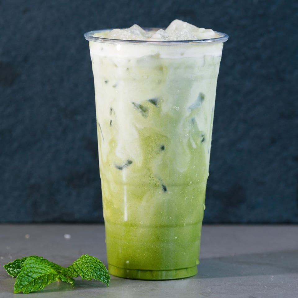 Iced green tea with milk and ice in a clear plastic cup, with mint leaves nearby.