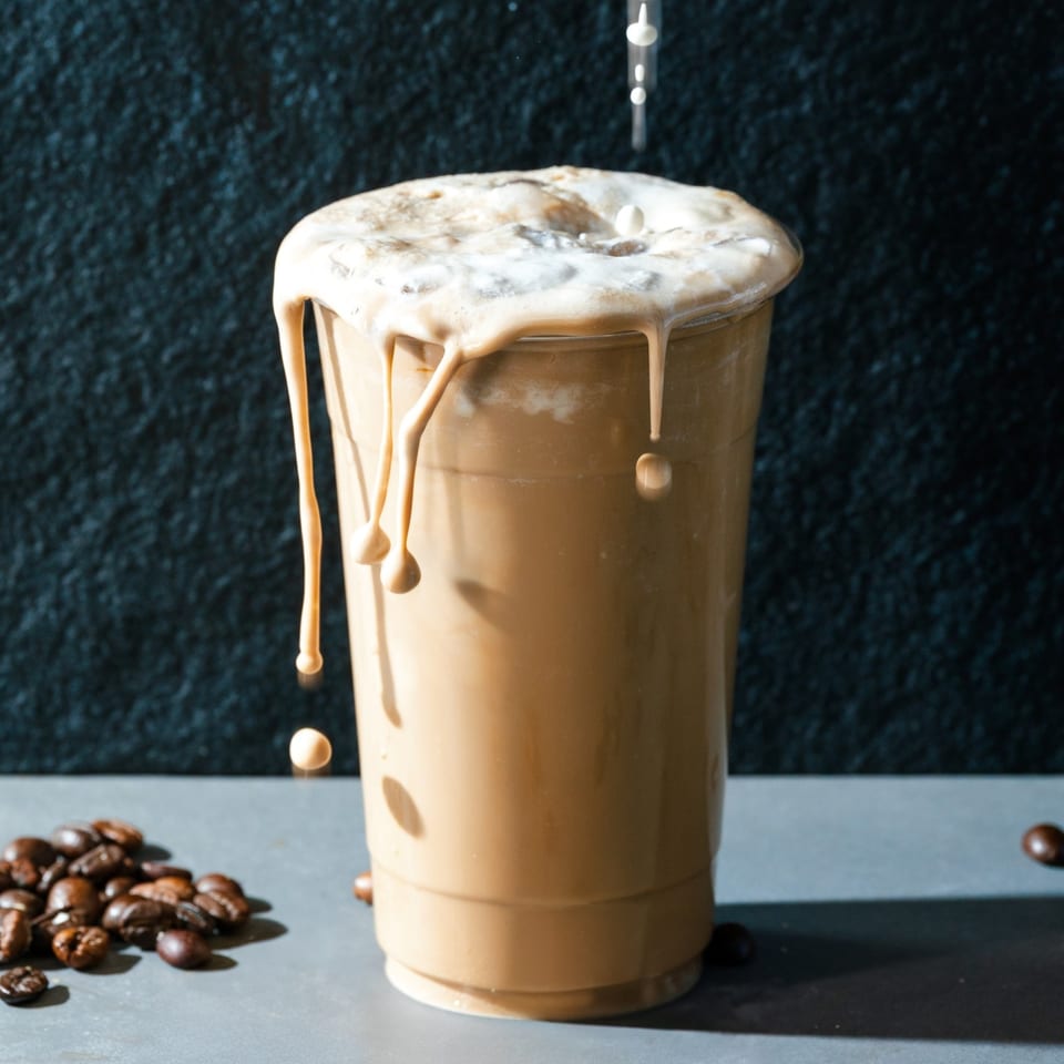 Iced Vietnamese coffee overflowing with foam, drips running down the cup, with coffee beans scattered nearby.