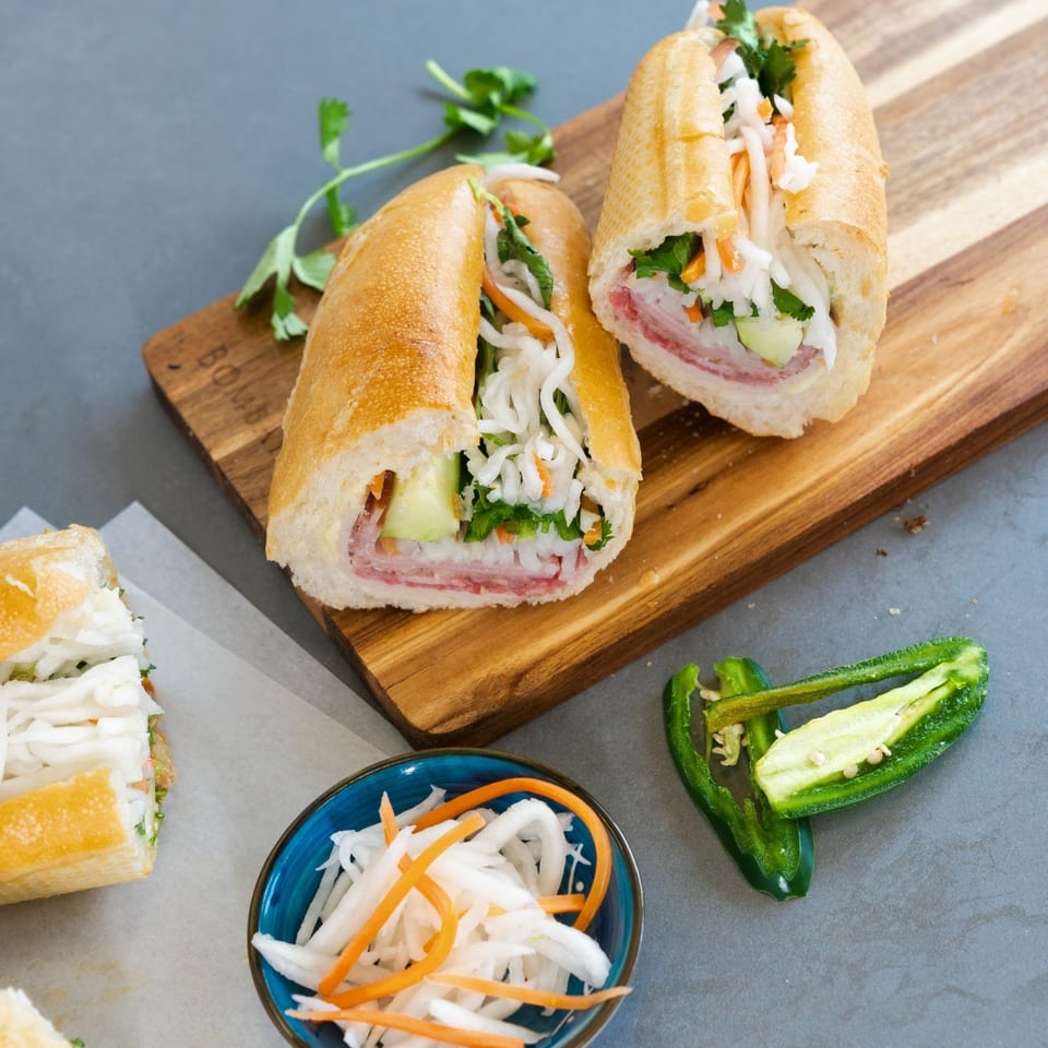 Two Saigon Blvd Bánh Mì sandwiches on a wooden board, with pickled vegetables and jalapeños.